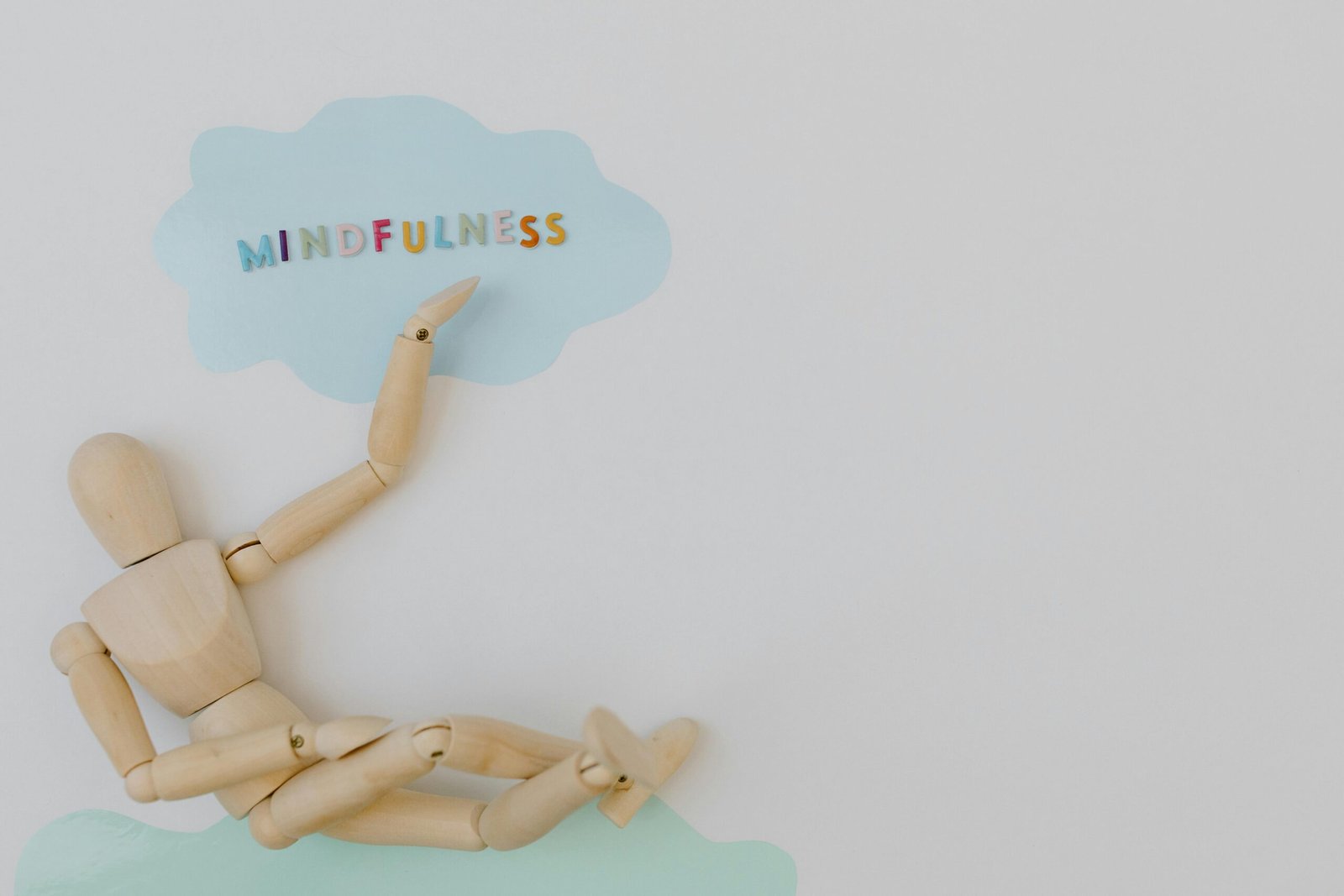 A wooden mannequin sitting in a relaxed pose on a white background, pointing toward a light blue cloud shape with the word “Mindfulness” written in colorful letters, symbolizing calm awareness and mental clarity