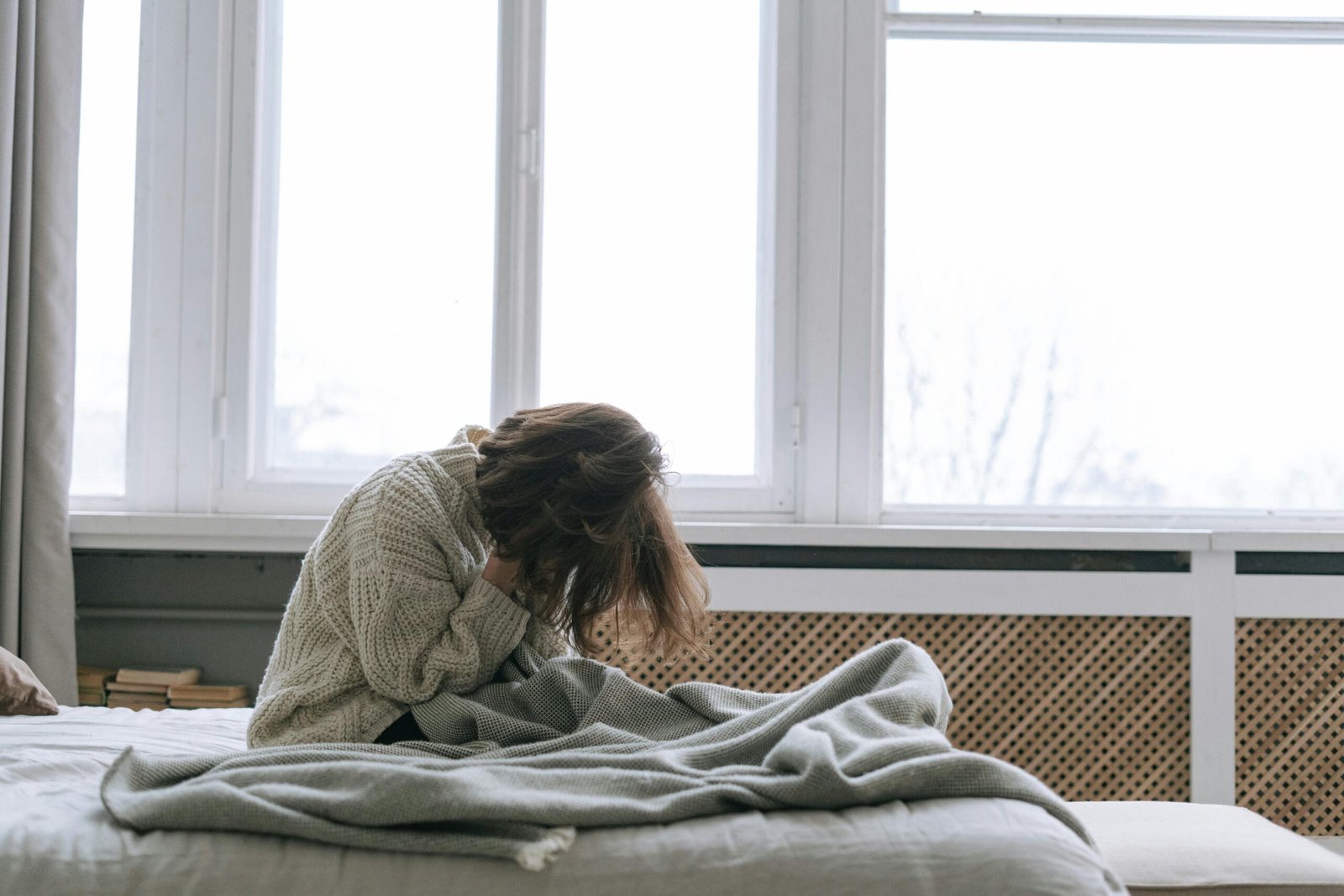 A stressed woman sitting on a bed with her head down, wrapped in a blanket near a window, showing signs of mental fatigue and daily stress.