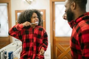 A child brushing their teeth in the bathroom while smiling at an adult beside them, both wearing matching red plaid pajamas during a morning routine