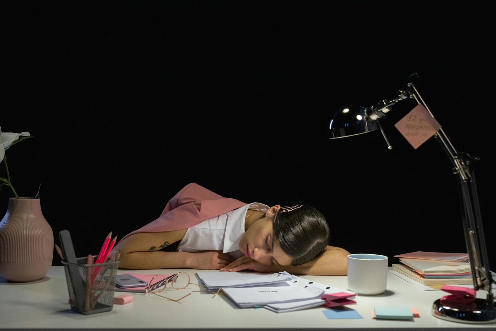 A tired woman asleep at her desk, resting her head on paperwork under a desk lamp, surrounded by office supplies, sticky notes, and a coffee mug, showing work fatigue and burnout